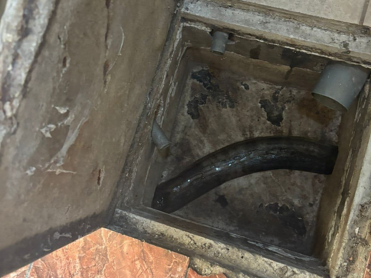 Cámara de inspección abierta en el piso de una casa, con la tapa de concreto levantada y una manguera negra adentro
